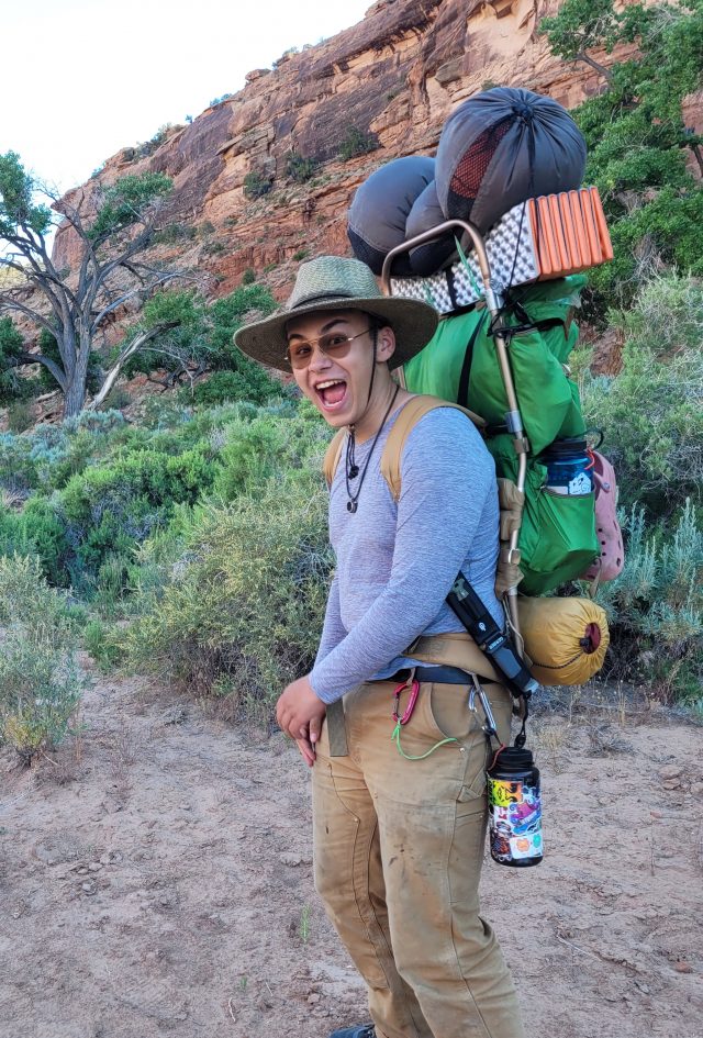 boy with rocks in the background smiling and carrying a big backpack