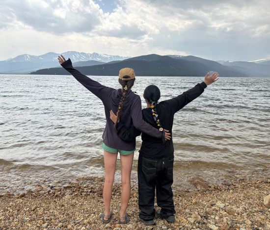 Two students stand near a lake with one arm around each other and the other outstretched toward the sky.